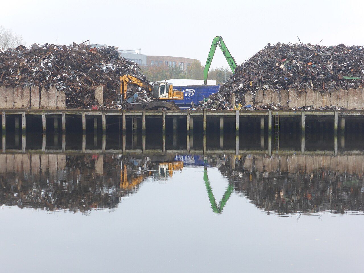 Mountains of scrap metal and a grab crane reflected in the still water of a dockside canal